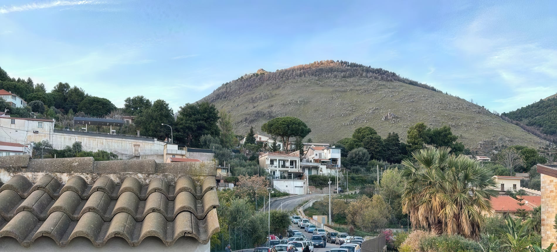 La strada che sale verso la sede di Monreale nel giorno della distribuzione, con la fila delle auto e le colline sullo sfondo